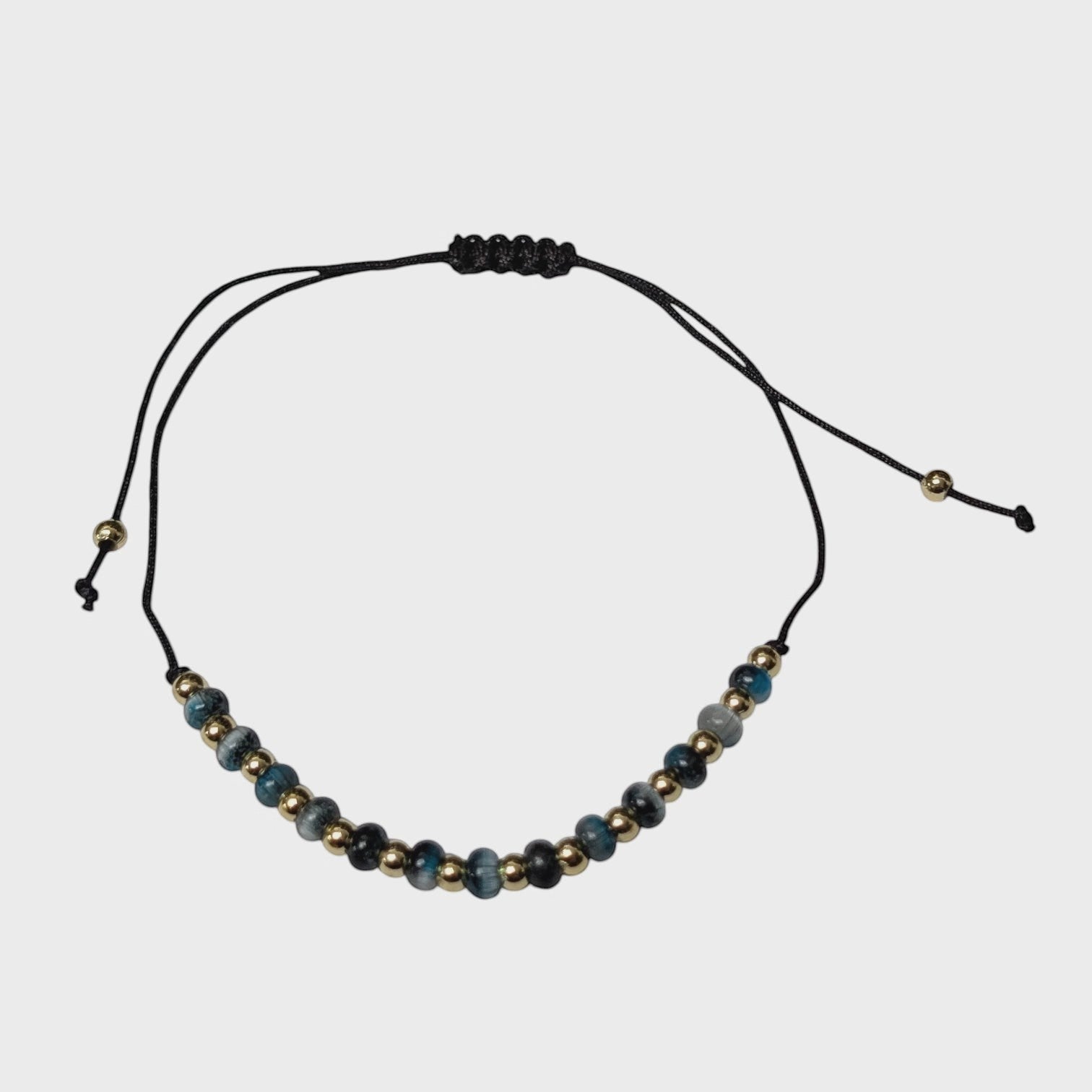 Aramat Jewels verstelbaar enkelbandje met blauwe cats eye kralen en gouden accenten op een zwart touwtje.